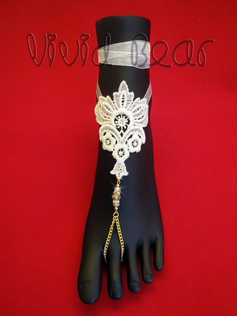 May include: White lace foot jewelry with a gold chain and crystal beads. The jewelry is on a black foot model against a red background.