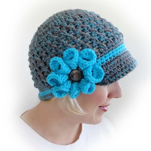 Puede incluir: Sombrero de punto gris y turquesa con un gran adorno de flor turquesa. El sombrero tiene una visera y est&aacute; hecho con un punto texturizado.