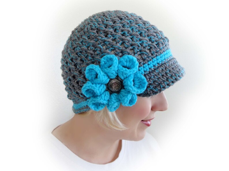 Puede incluir: Sombrero de punto gris y turquesa con un gran adorno de flor turquesa. El sombrero tiene una visera y est&aacute; hecho con un punto texturizado.