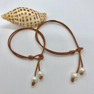 May include: Two brown leather bracelets with white pearl accents. The bracelets are tied with a knot and have a tassel-like design.