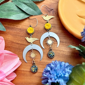 May include: A pair of celestial-themed earrings. Each earring features a silver crescent moon, a gold sun, and a small, round, colorful charm. Gold-colored bird and yellow stone accents add detail. The earrings hang from silver hooks.