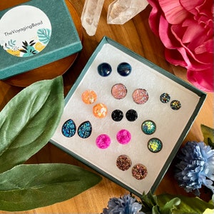 May include: A set of 12 pairs of earrings in a white box. The earrings are made of various materials, including glitter, resin, and metal. The earrings are in a variety of colors, including pink, blue, green, orange, and black.