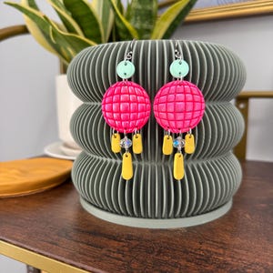May include: A pair of vibrant earrings featuring a light green disc, a hot pink sphere with a grid pattern, and yellow teardrop shapes. The earrings are displayed on a gray, ribbed decorative pot. The background includes a plant with green and yellow leaves.