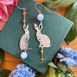 May include: A pair of silver rabbit silhouette earrings with blue agate beads and gold star charms. The earrings are hanging from a green box.