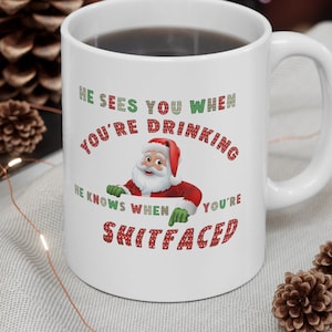 May include: White ceramic mug with a black liquid inside. The mug features a Santa Claus graphic and the text "HE SEES YOU WHEN YOU'RE DRINKING, HE KNOWS WHEN YOU'RE SHITFACED" in red and green lettering. Festive holiday theme.