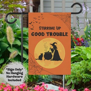 May include: Orange Halloween garden flag with the text "Stirring Up Good Trouble." The flag features a witch stirring a cauldron with a black cat, bats, and a spiderweb design. The flag is displayed on a black metal stand.