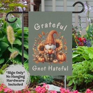 May include: A garden flag with a gnome design. The flag features a gnome holding a pumpkin, surrounded by sunflowers and pumpkins. The text reads "Grateful Gnot Hateful". The flag is displayed on a black metal stand.