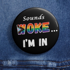 May include: A black button with the text "sounds WOKE... I'M IN" in white and rainbow colors. The button is round and has a glossy finish. The background is a blue denim fabric.