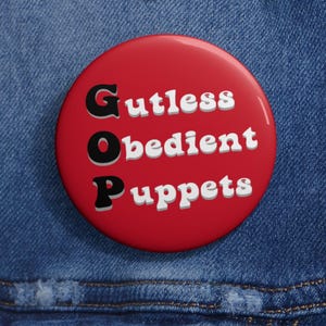 Puede incluir: Un botón rojo con el texto "Gutless, Obedient, Puppets" en letras negras y blancas. El botón está sobre un fondo de mezclilla azul.