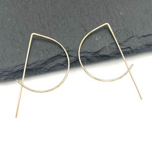 May include: A pair of gold-toned metal earrings featuring a geometric design. Each earring consists of a thin, continuous wire shaped into a sharp angular top point that transitions into a curved, open circular base with a straight tail end.