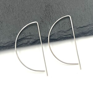May include: A pair of silver-toned earrings, each featuring a thin, curved wire forming a semi-circle shape with a straight vertical bar extending from the top, creating a clean, geometric design that is lightweight and simple in its construction.