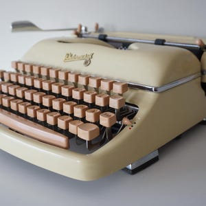 May include: A cream-coloured vintage typewriter with wooden keys and knobs. The typewriter has a curved design with a chrome trim and a silver carriage return lever. The keys are arranged in a standard QWERTY layout.