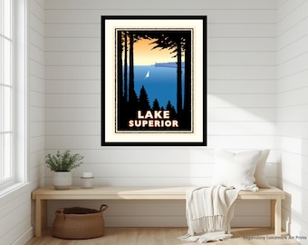 May include: A framed art print featuring a stylized landscape of Lake Superior. The image shows a blue lake with a small white sailboat, framed by black silhouettes of pine trees against a pale orange sky. The text LAKE SUPERIOR is printed in white.