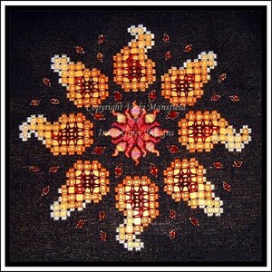 May include: A cross-stitch design featuring a circular pattern of eight stylized paisley shapes in shades of orange, red, and white on a black background. The center of the design features a stylized flower in shades of pink, red, and orange. The text "Copyright Vicki Mansfield In Grace Designs" is visible in the center of the design.
