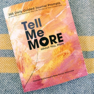 May include: A book titled "Tell Me More about You, Mom" with a pink, yellow, and orange abstract design. The book is a guided journal with 365 daily prompts to record and preserve the life stories of a loved one.