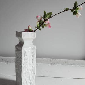 May include: A white ceramic vase with a textured surface holds a branch of pink flowers with green leaves.