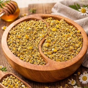 Peut inclure: Un bol en bois rempli de fleurs de camomille séchées, avec une cuillère en bois contenant plus de fleurs. Le texte "Chamomile Whole Flowers" est en haut de l'image. Les fleurs sont jaunes et vertes.