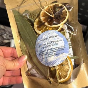 May include: A clear plastic bag filled with dried botanicals, including bay leaves and dehydrated lemon slices. The bag is tied with twine and features a label that reads "Heavenly Hollow's Simmer Pot Potpourri Breathe Easy."