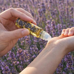 May include: A clear glass roller bottle filled with a yellow liquid and lavender buds is being applied to a wrist. The background is a field of purple lavender flowers. The bottle has a white rollerball applicator.