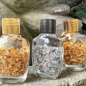 May include: Three glass bottles with gold and silver flakes inside. The bottles have gold, black, and gold lids.
