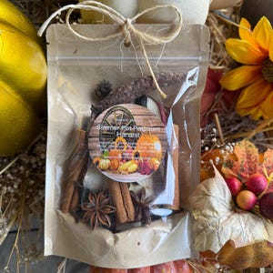 May include: A clear bag tied with twine, filled with potpourri ingredients. The bag features a circular label with the text "Simmer Pot Potpourri Harvest" and images of pumpkins and apples. Cinnamon sticks, star anise, and other dried botanicals are visible.