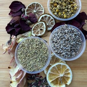 May include: Overhead shot of a wooden surface with various dried botanicals. Includes dried lemon slices, rose petals, chamomile flowers, lavender buds, and rosemary in small clear containers. The arrangement is colorful and natural.