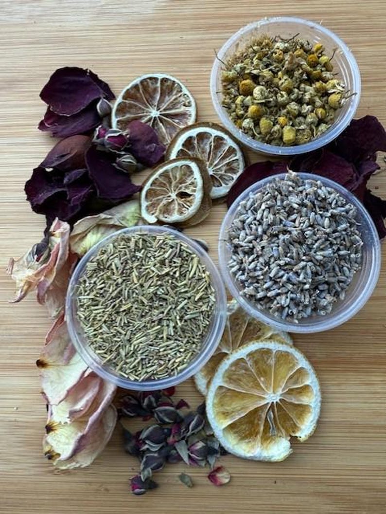 May include: Overhead shot of a wooden surface with various dried botanicals. Includes dried lemon slices, rose petals, chamomile flowers, lavender buds, and rosemary in small clear containers. The arrangement is colorful and natural.