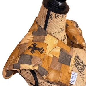 May include: A quilted oven mitt with a patchwork design in shades of brown, tan, and gray. A black cross is centered on the mitt. The mitt is displayed on a mannequin.