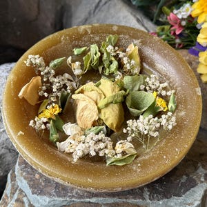 May include: A round, amber-colored candle holder filled with dried flowers and greenery. The arrangement includes yellow rose petals, small white flowers, and green leaves. The candle holder sits on a stone surface.