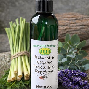 May include: A green glass spray bottle of Heavenly Hollow Natural & Organic Tick & Bug Repellent, 8 oz. The label features a red tick illustration. Bundles of lemongrass and lavender sprigs are in the background.
