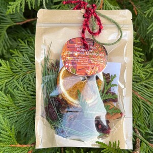 May include: A clear cellophane bag filled with potpourri, including dried orange slices, pine needles, and berries. A round tag with the text "Simmer Pot Potpourri Christmas Memories" is attached with red and green twine. The background is green foliage.