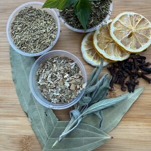May include: An overhead shot of various herbs and spices on a wooden surface. The image includes small plastic containers filled with dried herbs, fresh mint, dried lemon slices, cloves, and bay leaves. The colors are natural and earthy.