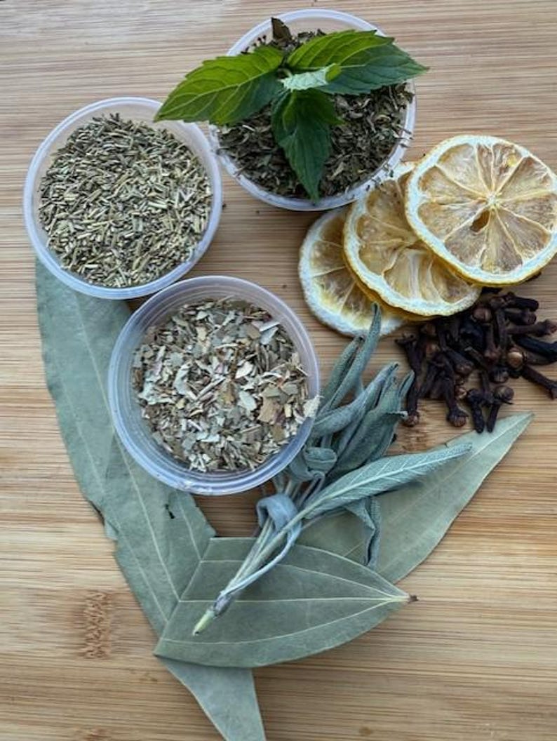 May include: An overhead shot of various herbs and spices on a wooden surface. The image includes small plastic containers filled with dried herbs, fresh mint, dried lemon slices, cloves, and bay leaves. The colors are natural and earthy.