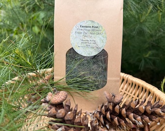 May include: A brown paper bag with a clear window, filled with fresh-cut Eastern Pine needles. A round label with text is affixed to the bag. Pine cones and pine needles are in a wicker basket in the foreground.