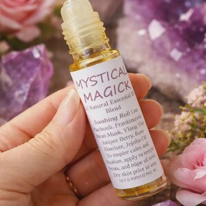May include: A clear glass roll-on bottle with a yellow liquid inside, labeled "MYSTICAL MAGICK." The label lists essential oils like Patchouli and Ylang Ylang. The bottle is held against a backdrop of flowers and crystals.