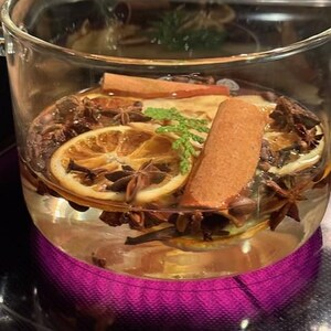 May include: A clear glass mug filled with a warm beverage, featuring slices of dried orange, cinnamon sticks, star anise, and sprigs of greenery. The mug sits on a purple heating element, suggesting a cozy, aromatic drink.