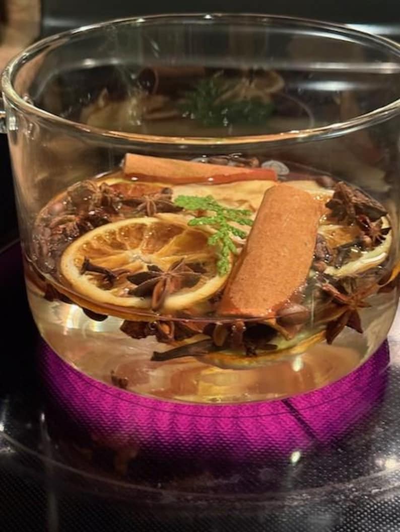 May include: A clear glass mug filled with a warm beverage, featuring slices of dried orange, cinnamon sticks, star anise, and sprigs of greenery. The mug sits on a purple heating element, suggesting a cozy, aromatic drink.