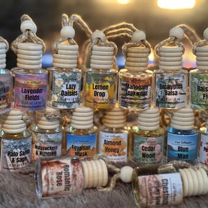 May include: A collection of 18 small glass bottles with wooden stoppers and twine hangers. Each bottle contains a car diffuser with various scents, including Pumpkin Harvest, Lavender Fields, Lazy Daisies, Lemon Drop, Mahogany Teakwood, Eucalyptus Balsa, Balsam Fir, Coffee Espresso, Palo Santo & Sage, Nutcrackers & Nutmeg, Egyptian Musk, Almond Honey, Cedar Wood, Blue Man, Peppermint Candy, Romantic Rose, and Chestnut.