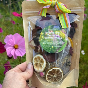 May include: A clear bag of potpourri with dried flowers, herbs, and citrus slices, tied with a colorful striped ribbon. The bag has a label that reads "Simmer Pot Potpourri Summer Vibes". The background is a garden with pink flowers.