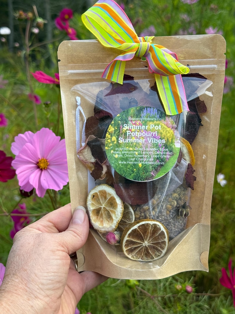 May include: A clear bag of potpourri with dried flowers, herbs, and citrus slices, tied with a colorful striped ribbon. The bag has a label that reads "Simmer Pot Potpourri Summer Vibes". The background is a garden with pink flowers.