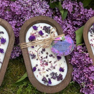 May include: Three wooden bowl candles with white wax, decorated with purple flowers and crystals. The candles are surrounded by purple lilac flowers. One candle has a tag that reads "Lilac Soy Candle Bowl".