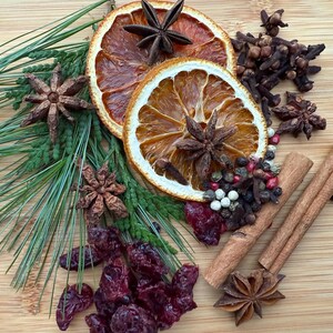 May include: A collection of dried orange slices, star anise, cloves, cinnamon sticks, cranberries, and peppercorns arranged on a wooden surface. Green pine needles add a touch of freshness to the arrangement. Aromatic spices for the holidays.