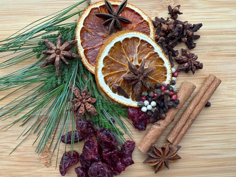 May include: A collection of dried orange slices, star anise, cloves, cinnamon sticks, cranberries, and peppercorns arranged on a wooden surface. Green pine needles add a touch of freshness to the arrangement. Aromatic spices for the holidays.