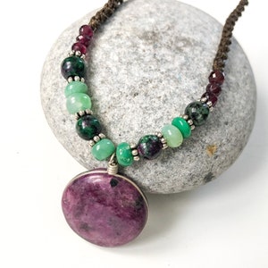 May include: A necklace with a large, round, purple pendant and green and burgundy beads. The necklace is made with a brown cord and silver accents.