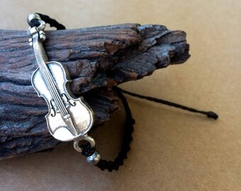 May include: A black cord bracelet with a silver violin charm. The bracelet is on a piece of driftwood.
