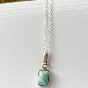 Op de afbeelding: Een zilveren ketting met een rechthoekige, lichtblauwe hanger. De hanger heeft een gemarmerd uiterlijk en hangt aan een fijne zilveren ketting. De ketting wordt gepresenteerd op een witte ondergrond.