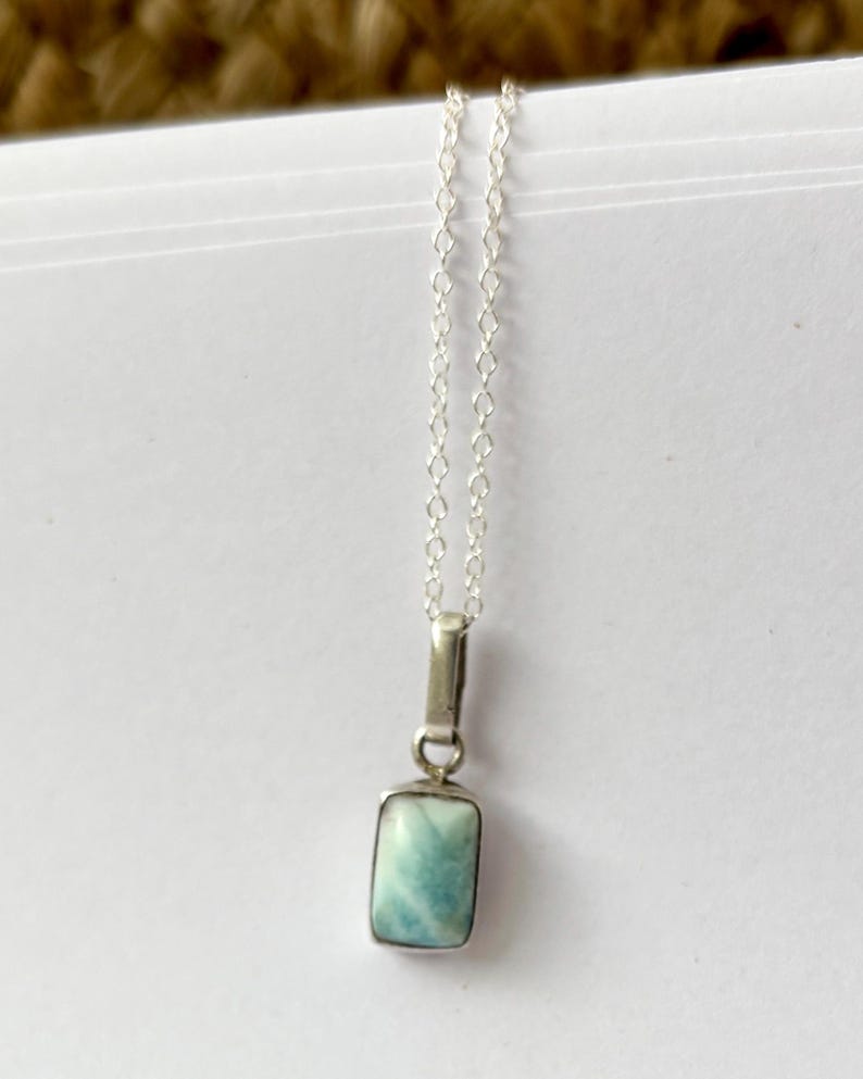 Op de afbeelding: Een zilveren ketting met een rechthoekige, lichtblauwe hanger. De hanger heeft een gemarmerd uiterlijk en hangt aan een fijne zilveren ketting. De ketting wordt gepresenteerd op een witte ondergrond.