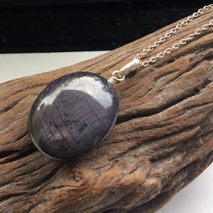 Puede incluir: Un collar de cadena de plata con un colgante ovalado azul oscuro. El colgante tiene un acabado brillante e iridiscente.