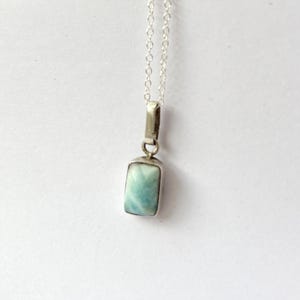 Op de afbeelding: Een rechthoekige hangerketting met een lichtblauwe edelsteen gezet in een zilverkleurig metalen frame. De hanger hangt aan een fijne zilveren ketting. De edelsteen heeft een gemarmerd uiterlijk met tinten blauw en wit.
