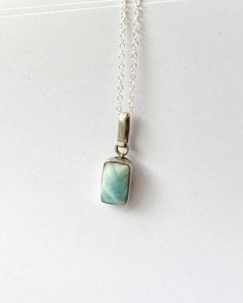 Op de afbeelding: Een rechthoekige hangerketting met een lichtblauwe edelsteen gezet in een zilverkleurig metalen frame. De hanger hangt aan een fijne zilveren ketting. De edelsteen heeft een gemarmerd uiterlijk met tinten blauw en wit.